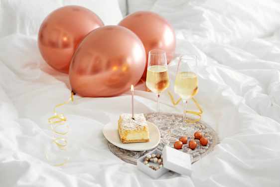Pink balloons, cake, sparkling wine glasses, present and chocolate on tray in bed